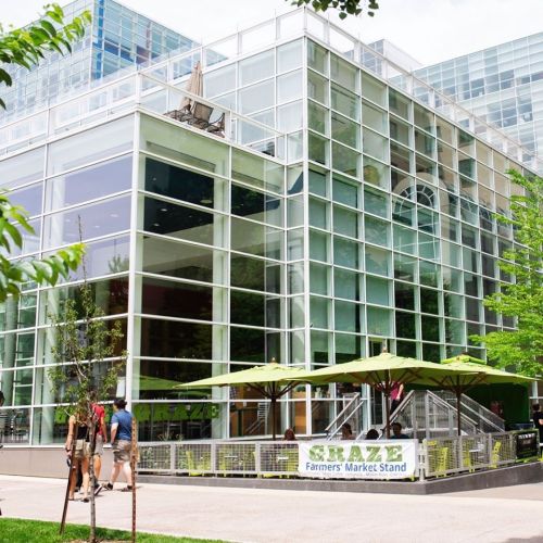 A modern glass building with outdoor seating and umbrellas, featuring a sign for a farmer's market stand.