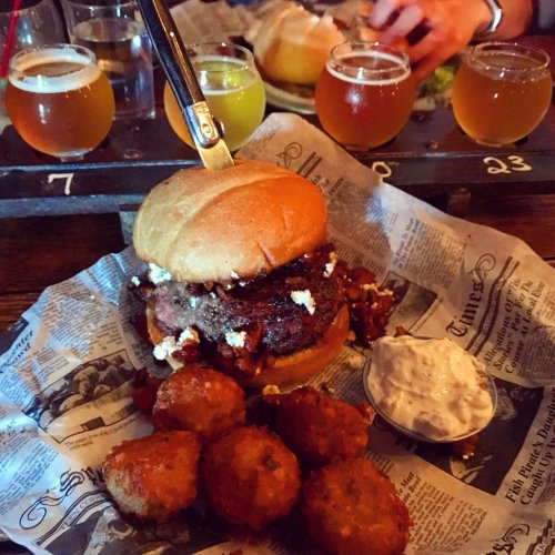 A hearty burger topped with cheese on a plate with fried bites and a dipping sauce, alongside a flight of craft beers.