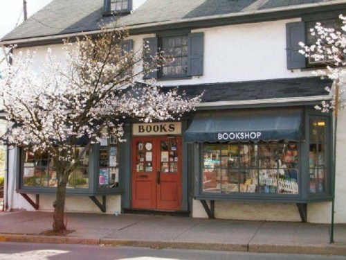 A charming bookshop with a red door, surrounded by blooming trees and large windows displaying books.