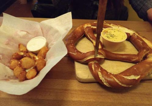A large pretzel on a wooden board with cheese dip, alongside a basket of tater tots and dipping sauce.