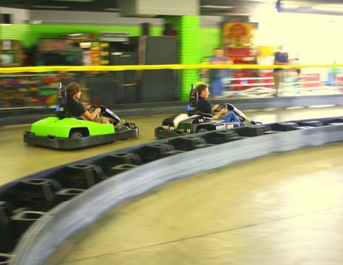 Two go-karts racing on a track, one green and one black, with a blurred background of an indoor amusement area.