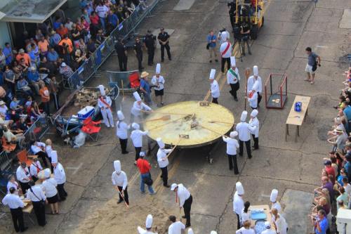 Chefs in white hats prepare a giant pizza on a large table, surrounded by a crowd at an outdoor event.