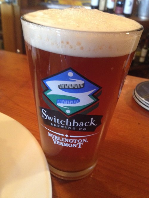 A glass of amber beer with a frothy head, featuring a Switchback Brewing Co. logo from Burlington, Vermont.