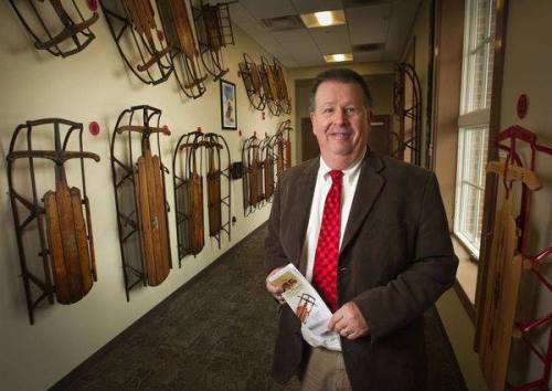 A man in a brown suit and red tie stands in a room lined with wooden sleds, holding a brochure.