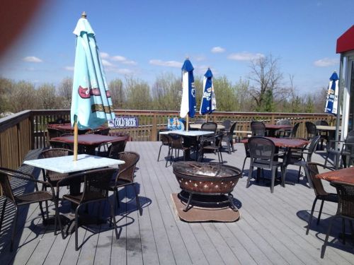 Outdoor patio with tables, chairs, and a fire pit, surrounded by trees and blue skies. Umbrellas provide shade.