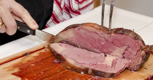 A person slices a large piece of cooked meat on a wooden cutting board, with juices visible on the surface.