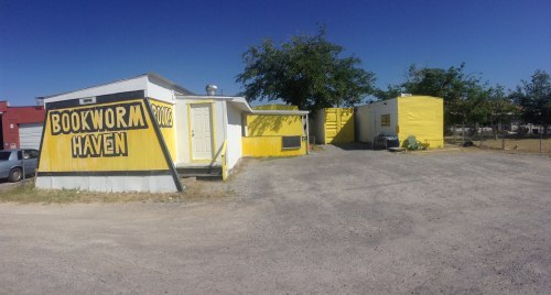 A bright yellow building with "Bookworm Haven" painted on the side, surrounded by gravel and trees under a clear blue sky.