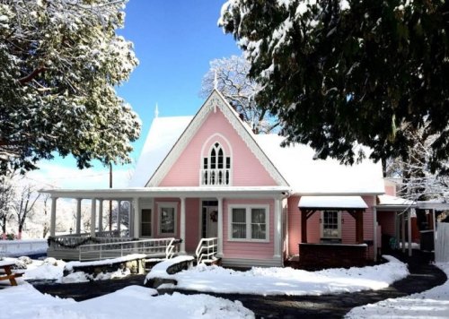 A charming pink house covered in snow, surrounded by trees and a clear blue sky.