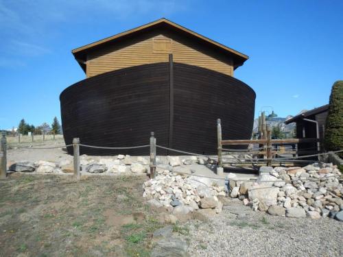 A large wooden structure resembling a boat, set in a gravel area with rocks and a fence, under a clear blue sky.