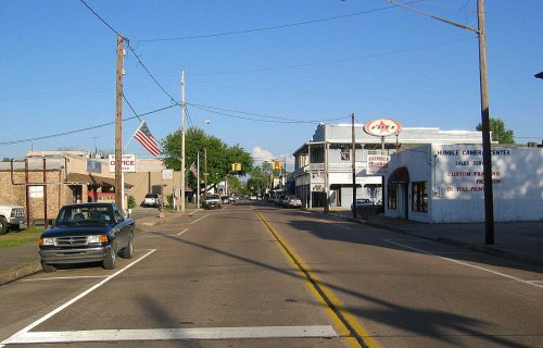 A quiet street scene with shops, a parked truck, and an American flag in the background under a clear blue sky.