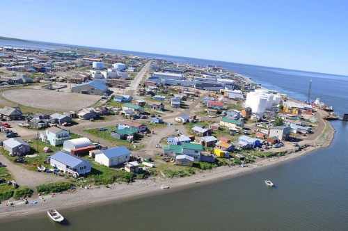 Aerial view of a coastal town with colorful buildings, roads, and boats along the shore under a clear blue sky.