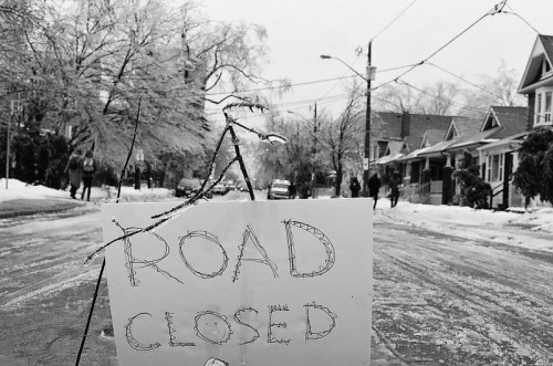 A handwritten "ROAD CLOSED" sign stands in a snowy street lined with houses and icy trees.