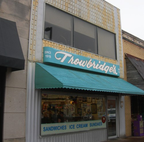 A vintage storefront with a teal awning, displaying "Throwbridge's" and offering sandwiches, ice cream, and sundaes.