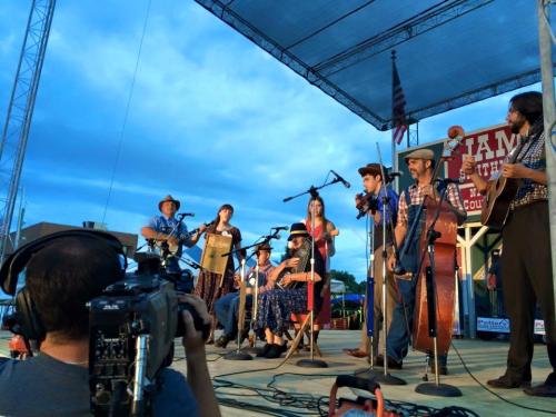 A bluegrass band performs on stage with various instruments, while a camera captures the scene under a cloudy sky.