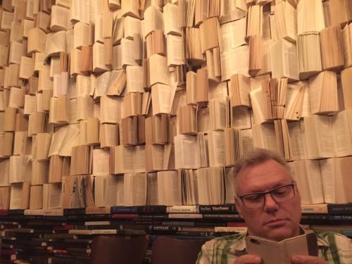 A man sits in front of a wall covered with open books, focused on his phone in a cozy, book-filled environment.