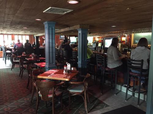 Interior of a cozy restaurant with wooden decor, tables, and patrons seated at the bar. Soft lighting creates a warm atmosphere.