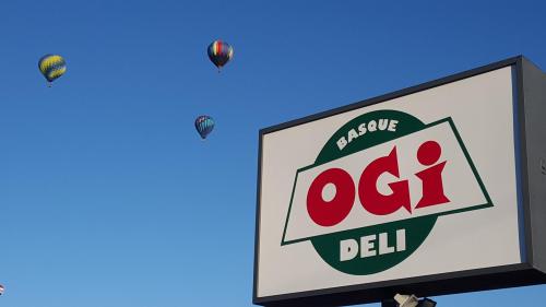 A sign for Ogi Deli with colorful hot air balloons in a clear blue sky above.