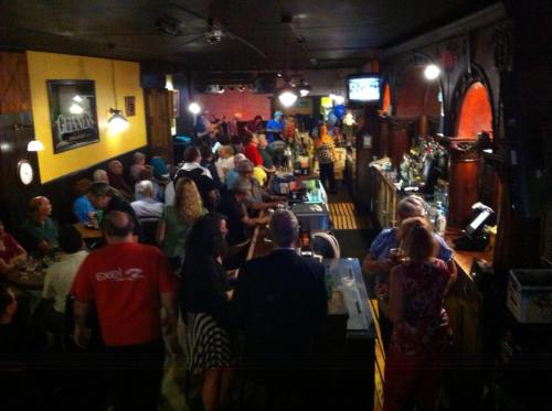 A lively bar scene with a crowd of people socializing, drinks at the counter, and colorful decorations in the background.