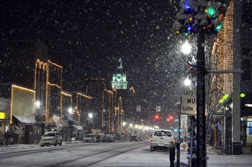 A snowy street at night, lined with festive lights and a clock tower in the background, creating a winter wonderland scene.