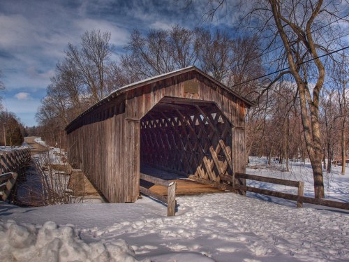 A rustic covered bridge surrounded by snow and trees under a clear blue sky.