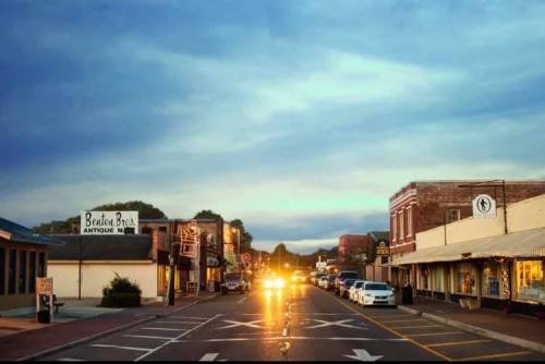 A quaint street scene at dusk, featuring shops and parked cars under a cloudy sky. Warm lights illuminate the road.