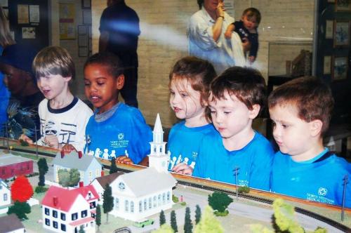 A group of children in blue shirts observe a model train display, smiling and engaged.
