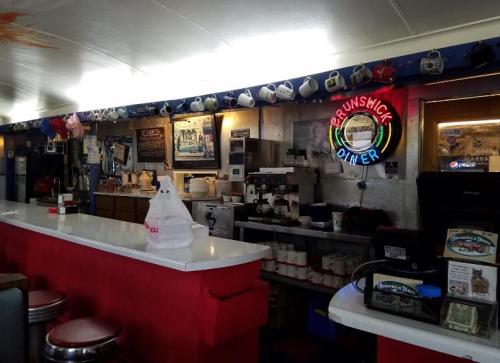 Interior of a diner featuring a counter, vintage decor, a neon sign, and a display of colorful coffee mugs.