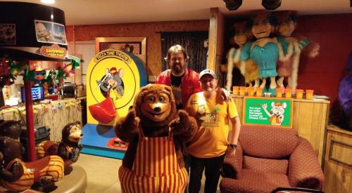 A man and a woman pose with a costumed bear character in a colorful, themed room with various decorations.