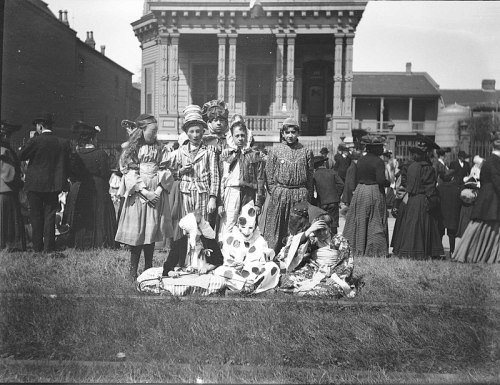 A group of children in traditional costumes poses in front of a historic building, with a crowd in the background.