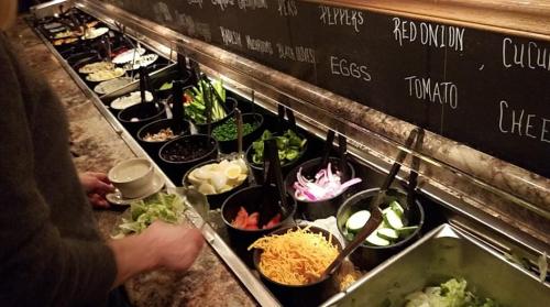 A salad bar with various toppings including vegetables, cheese, and eggs, with a person preparing a salad.
