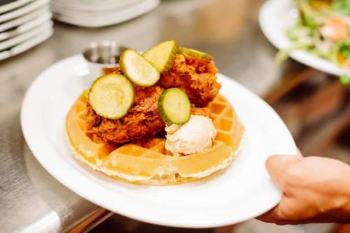 A plate of chicken and waffles topped with pickles and a scoop of ice cream, served on a white dish.