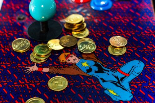 Close-up of an arcade game featuring a superhero design, with gold coins scattered on the colorful surface.