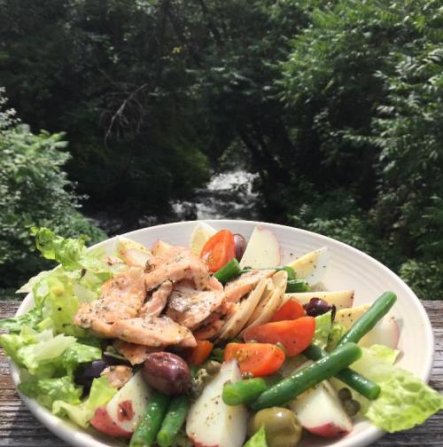 A vibrant salad with grilled salmon, mixed greens, cherry tomatoes, potatoes, olives, and green beans, set outdoors by a stream.