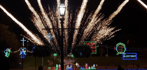 Colorful fireworks burst over a festive display of holiday lights, including angels, a cross, and a "Silent Night" sign.