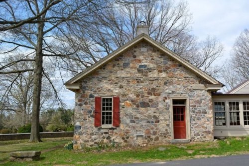 A stone house with a red door and shutters, surrounded by trees and a clear blue sky.