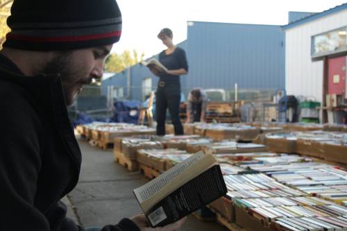 A man sits on the ground reading a book, while a woman stands nearby reading among stacks of books outdoors.