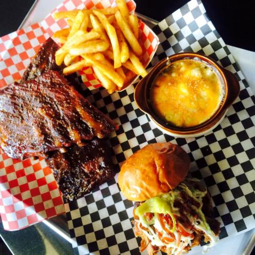 A plate featuring BBQ ribs, seasoned fries, cheesy baked dish, and a pulled pork sandwich on a checkered paper.