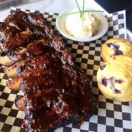 A plate of glazed ribs with mashed potatoes and blueberry muffins on a checkered paper background.