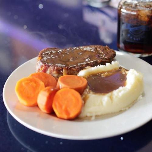 A plate of meatloaf with gravy, mashed potatoes, and steamed carrots, with a glass of cola in the background.