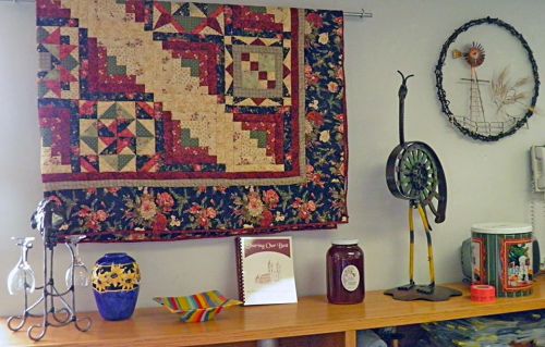 A colorful quilt hangs on the wall above a wooden shelf with decorative items, including vases and a book.