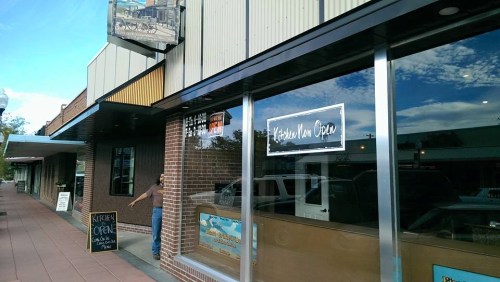 A storefront with a sign reading "Kitchen Now Open" and a person walking out, under a blue sky with clouds.