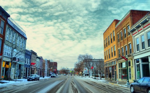 A snowy street lined with historic buildings and shops under a cloudy sky. Cars parked along the road.