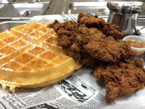 A plate featuring crispy fried chicken, a golden waffle, and a small cup of syrup on a newspaper-lined tray.