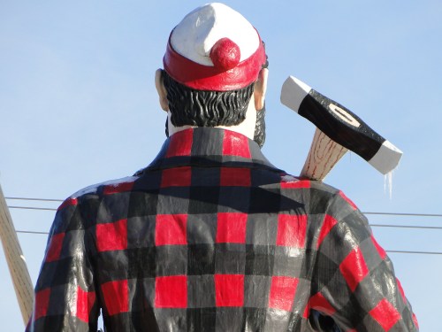 A large statue of a lumberjack in a red and black checkered shirt, holding an axe, viewed from behind against a blue sky.