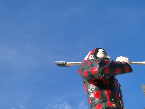 A large statue of a lumberjack in a red and black checkered shirt, holding a spear against a clear blue sky.