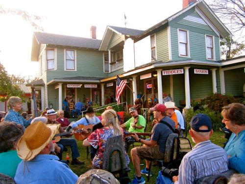 A group of musicians and spectators gather outside a green house, playing instruments and enjoying a lively jam session.