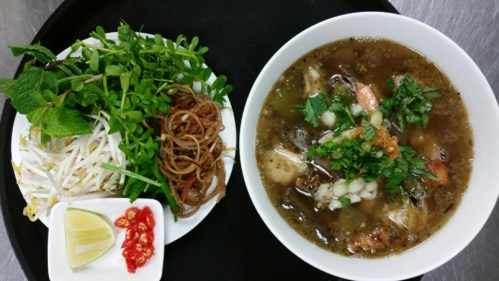 A bowl of soup with shrimp and herbs, accompanied by a plate of fresh greens, bean sprouts, and lime wedges.