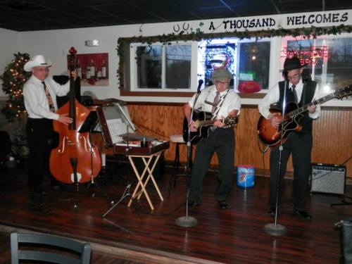 Three musicians perform in a cozy venue, playing guitar and double bass, with festive decorations in the background.