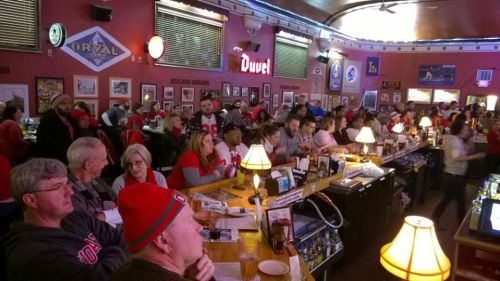 A crowded bar with patrons watching a game, wearing red attire, and sitting at the bar and tables. Warm lighting throughout.