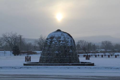 A snow-covered landscape features a large stone structure under a hazy sun, with trees and a fence in the background.
